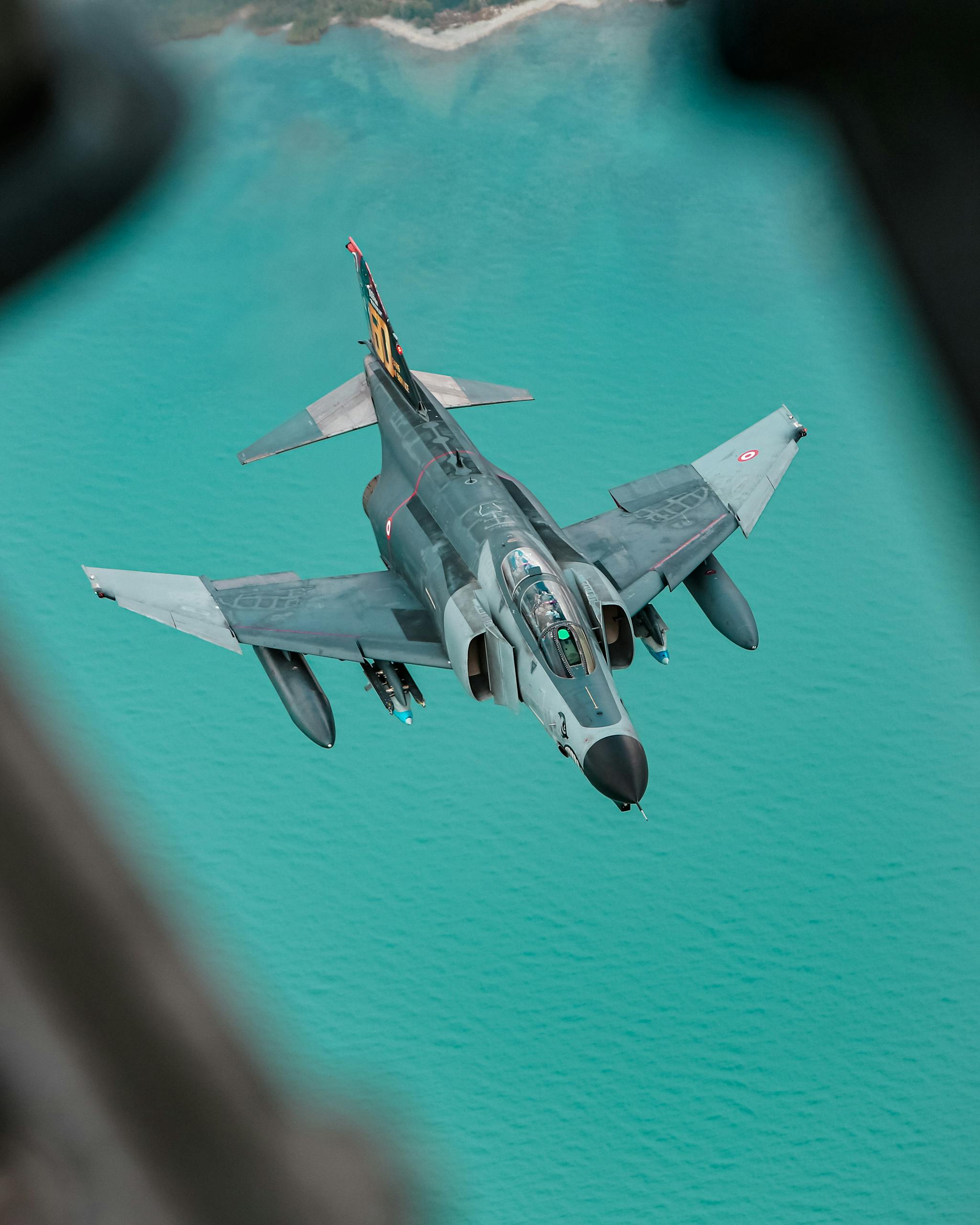 Aerial view of a military fighter jet soaring above vibrant turquoise waters.