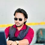 Man with sunglasses working intently on a laptop indoors. Casual professional attire.