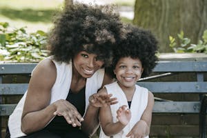A joyful mother and daughter with afro hair smiling together on a park bench.