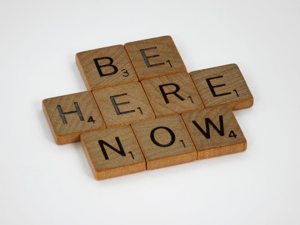 Inspirational text 'Be Here Now' created with wooden Scrabble tiles on white background.