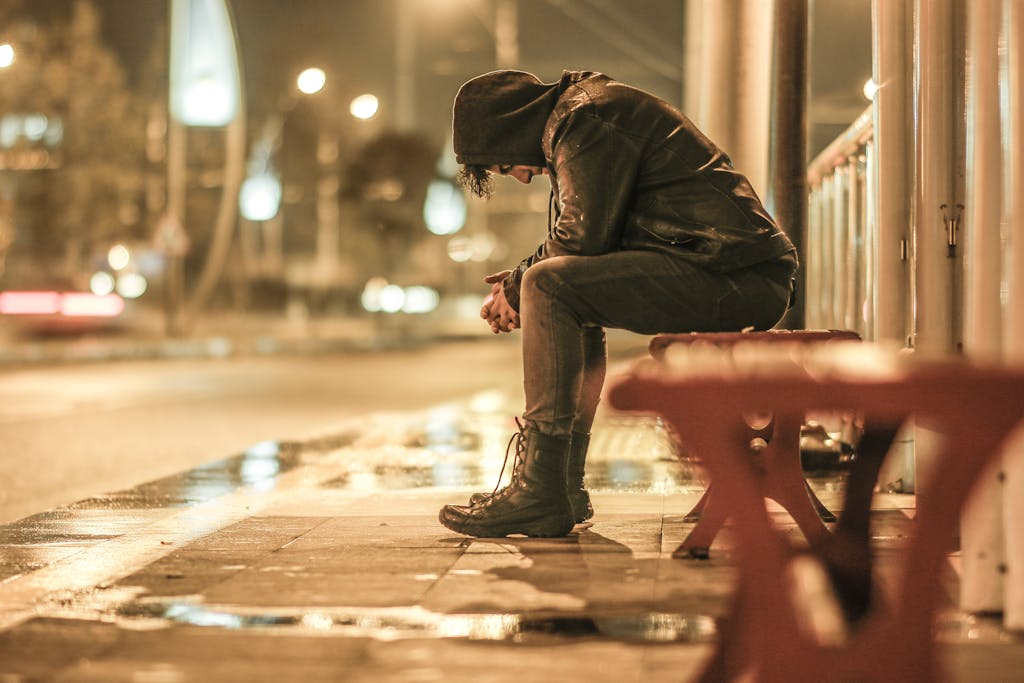Man in hoodie sitting on bench at night, reflecting in rainy urban setting.
